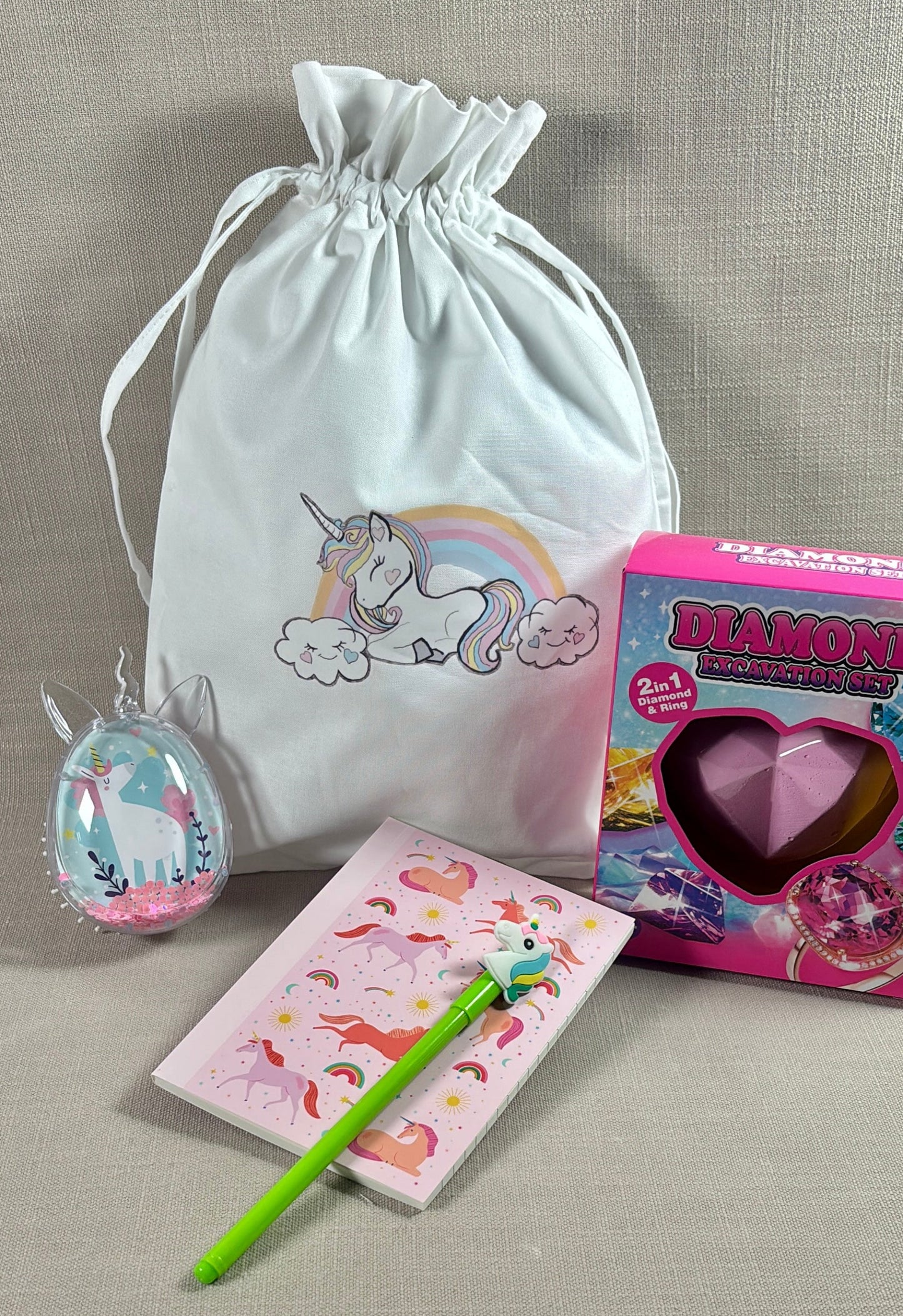 A unicorn-themed gift set on a beige fabric background, including a white drawstring bag with a unicorn print, a pink heart-shaped chocolate box, a small unicorn ornament, a pink notebook with unicorn illustrations, and a green pen.