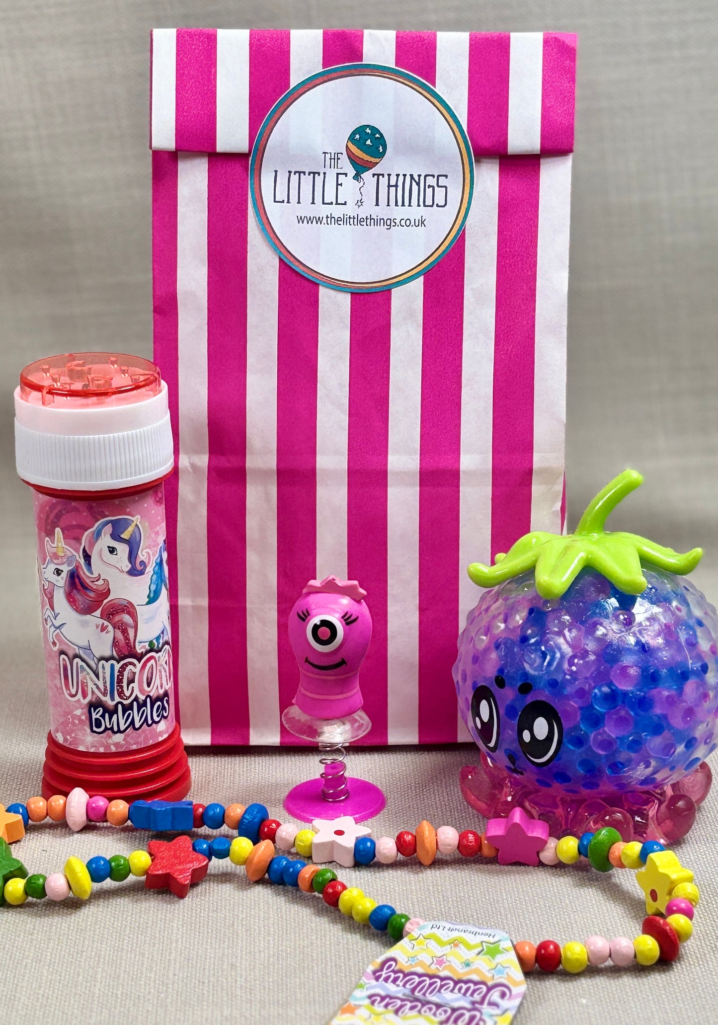 Children's toys, including a squishy toy, wooden necklace, Bubbles, and a pop up monsters, with a striped paper gift bag featuring 'The Little Things' sticker.