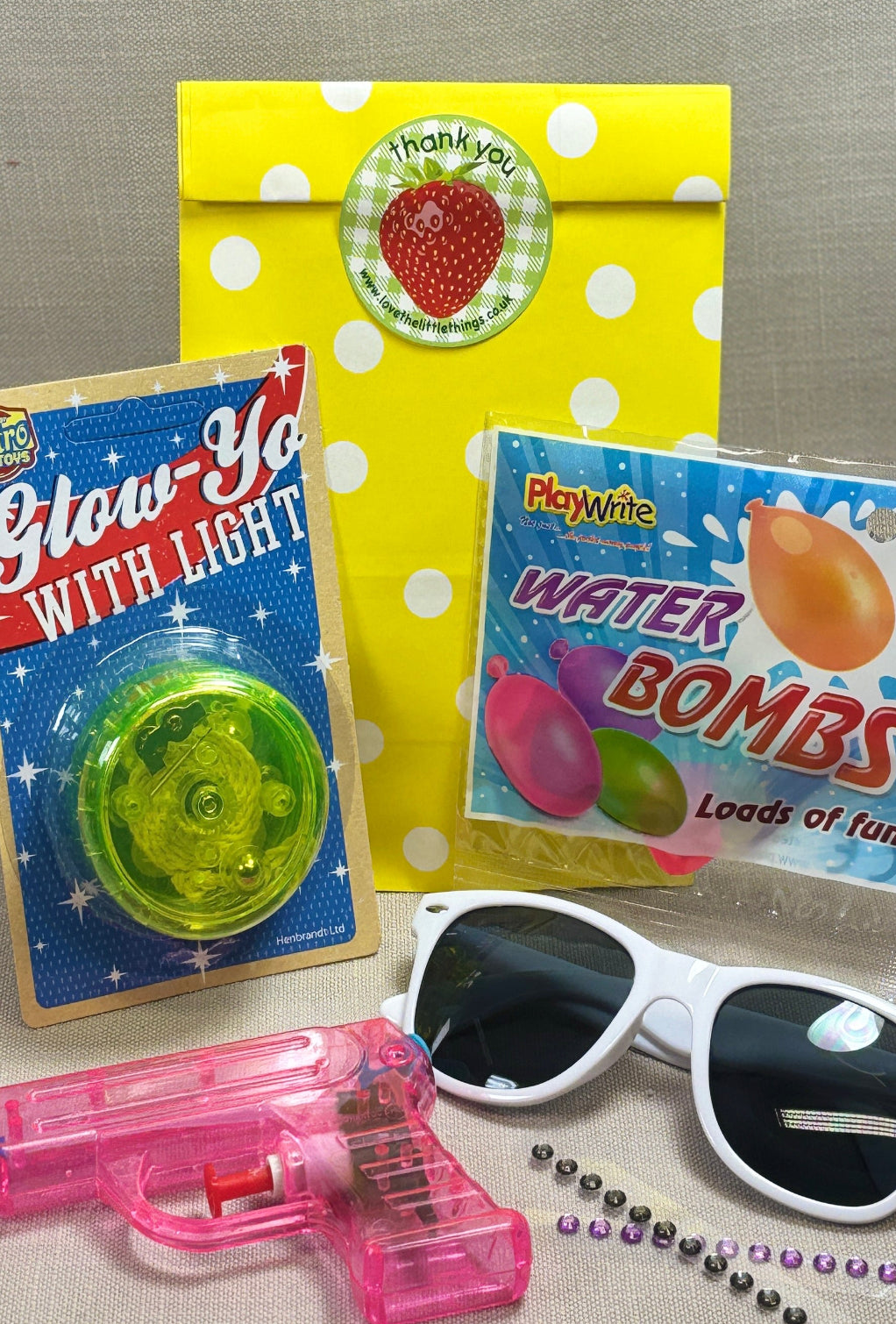 Yellow with white spots paper party bag with a strawberry sticker to seal. Included in the bag is a water pistol, wanter bombs, yoyo and sunglasses for kids.
