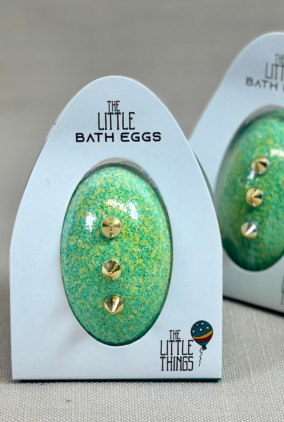 Two green bath eggs with gold accents in their packaging on a beige background