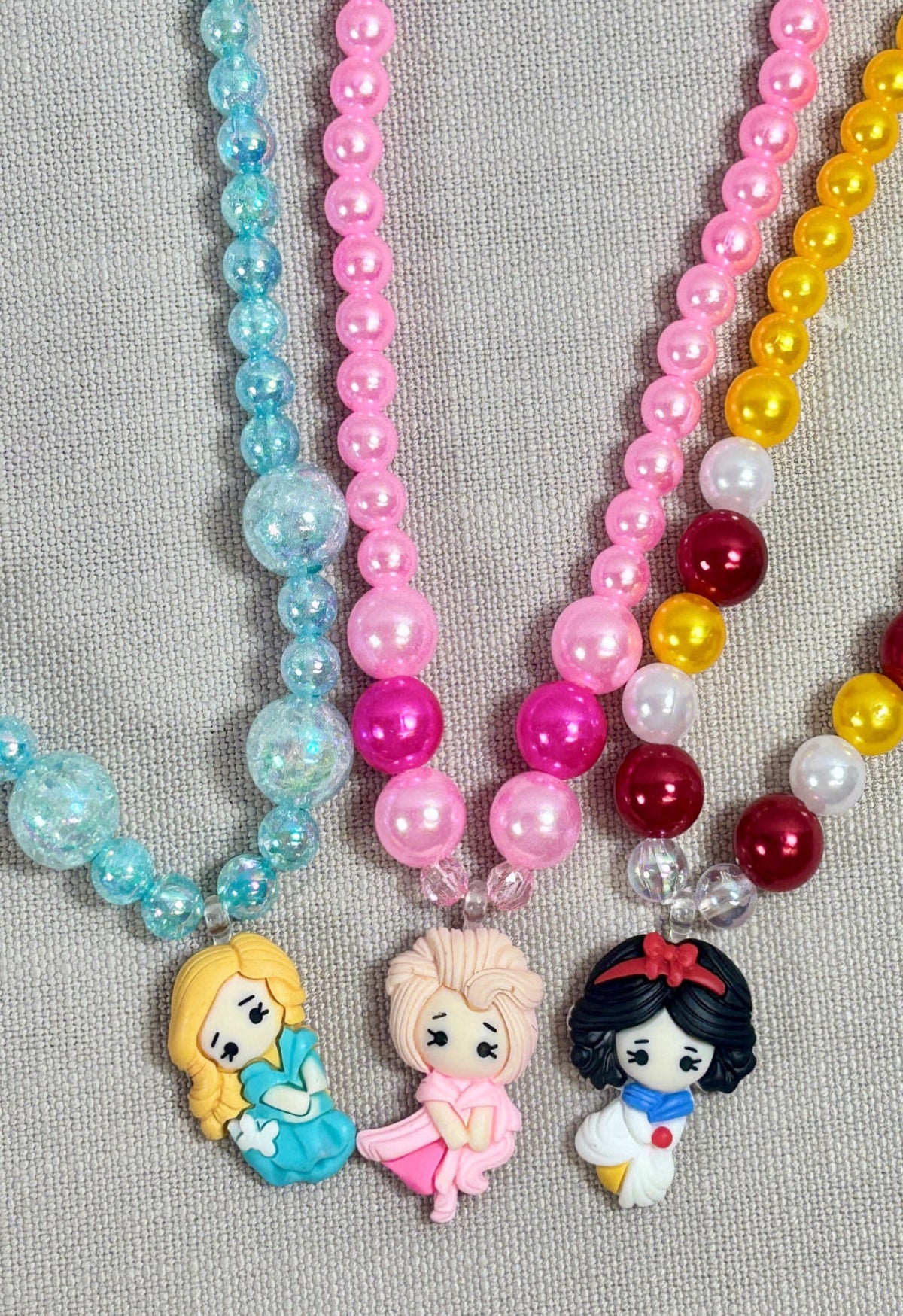 Three colorful beaded necklaces with cartoon princess character pendants on a gray background