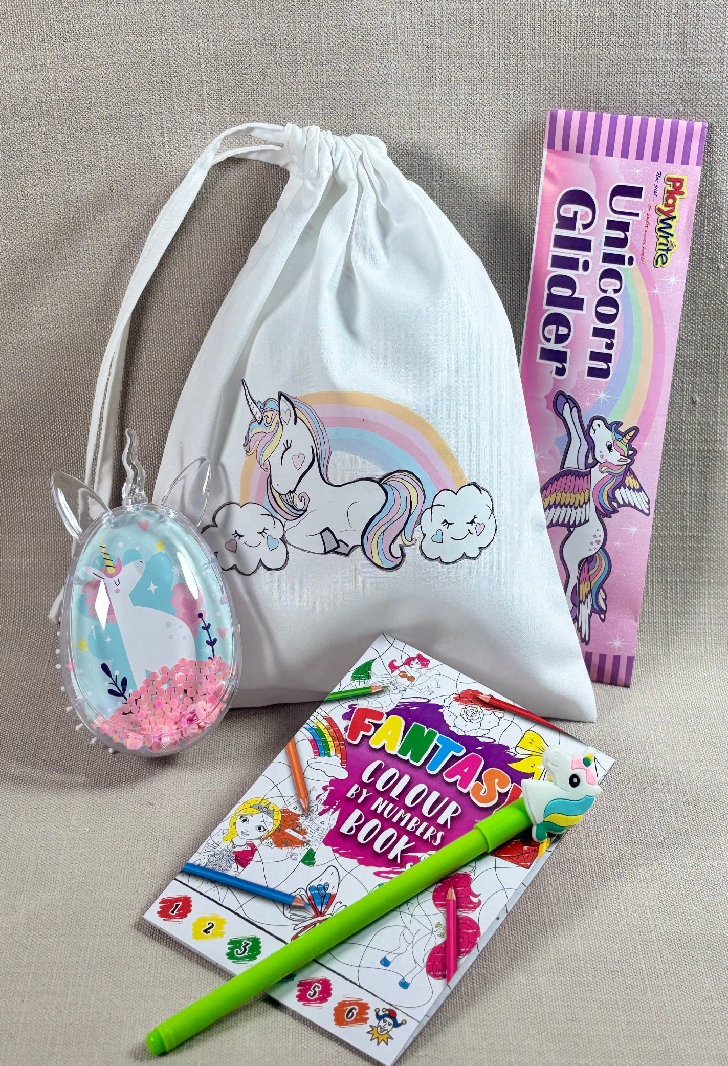 A white drawstring pouch with a unicorn and rainbow print, shown with a pink 'Unicorn Glider' book, a 'Fantasy Colouring Book', and a green pen on a beige textured fabric background.