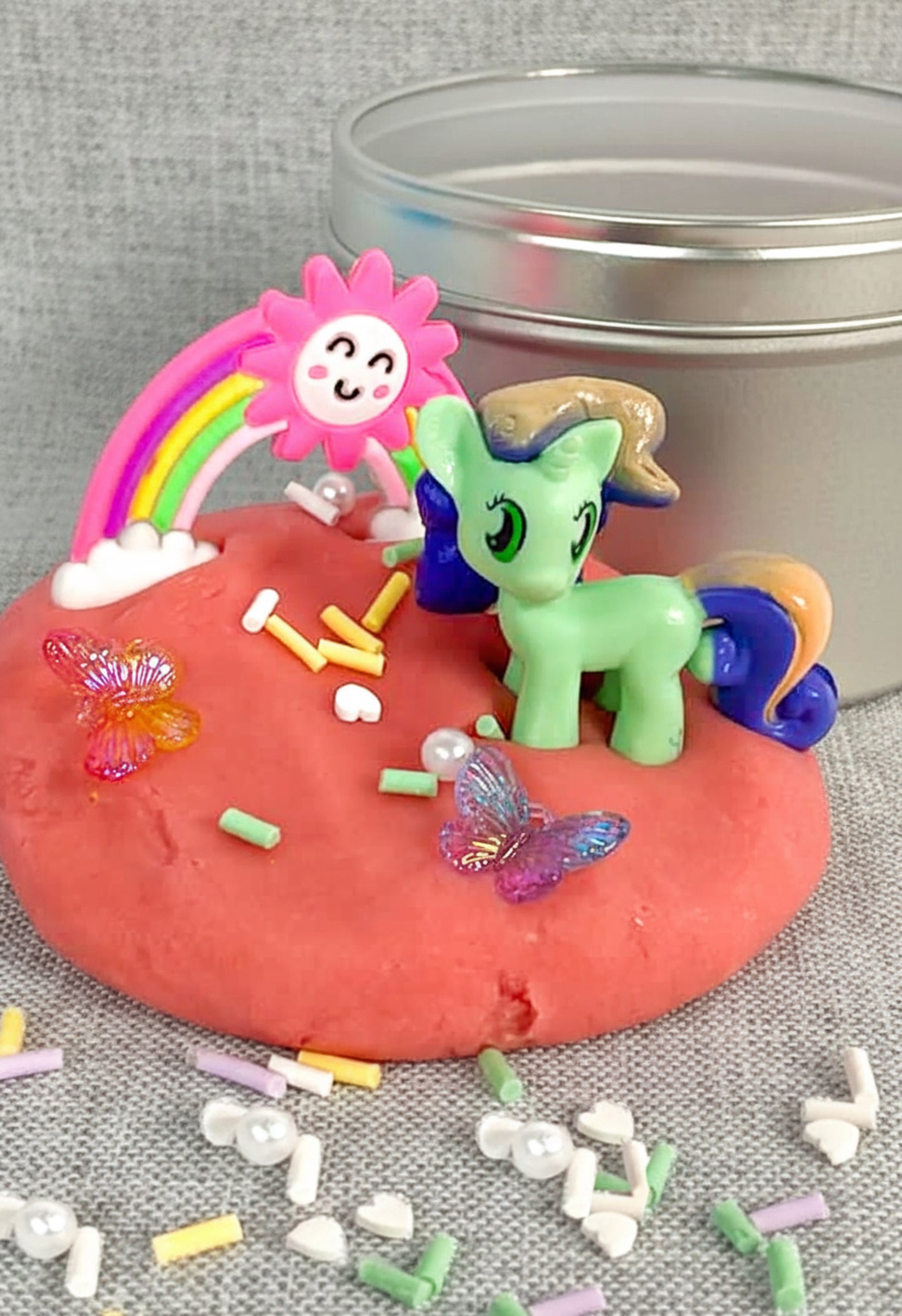 A play dough kit with various colorful themed shapes and toys spilling out, including a green unicorn figurine and small plastic objects on a red surface inside a metallic tin.