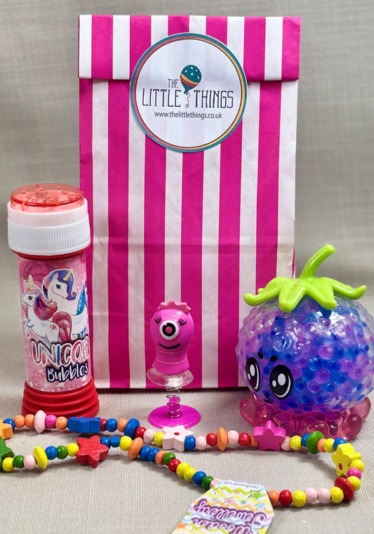 Children's toys, including a squishy toy, wooden necklace, Bubbles, and a pop up monsters,  with a striped paper gift bag featuring 'The Little Things' sticker.