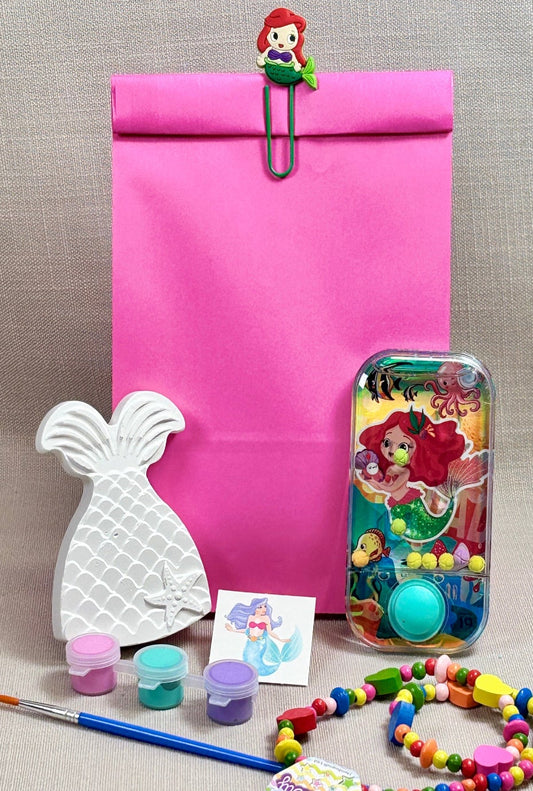 Pink paper party bag with mermaid paper clip. Mermaid tail ceramic with paint and a paint brush. a mermaid water game and a wooden beaded necklace.
