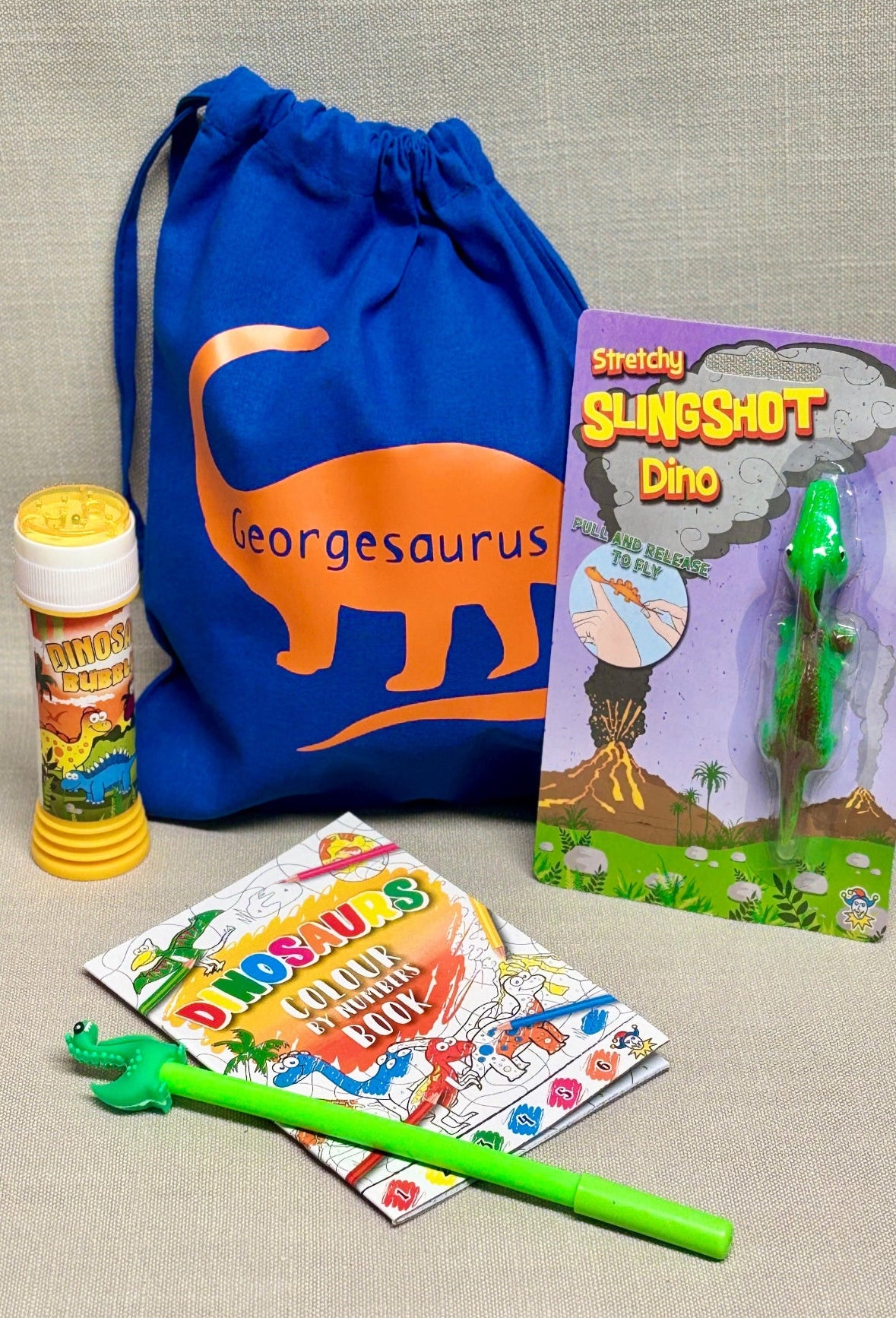 Dinosaur-themed children's items including a blue drawstring bag, Slingshot Dino toy, and activity book on a beige surface.