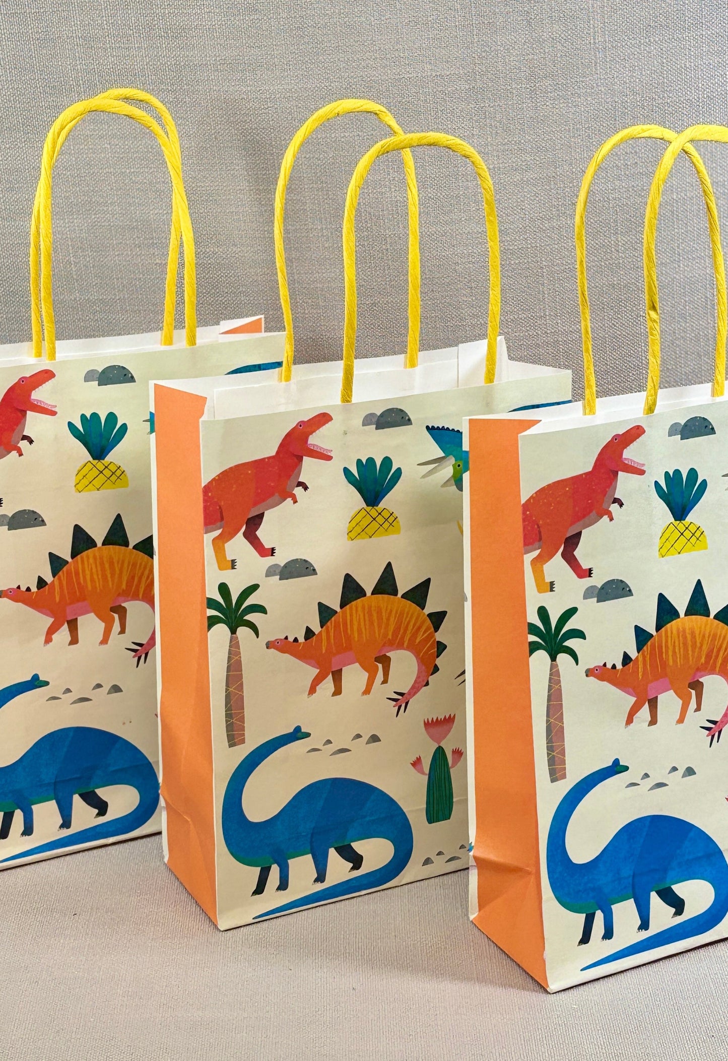Three dinosaur-themed paper bags with yellow handles on a gray background