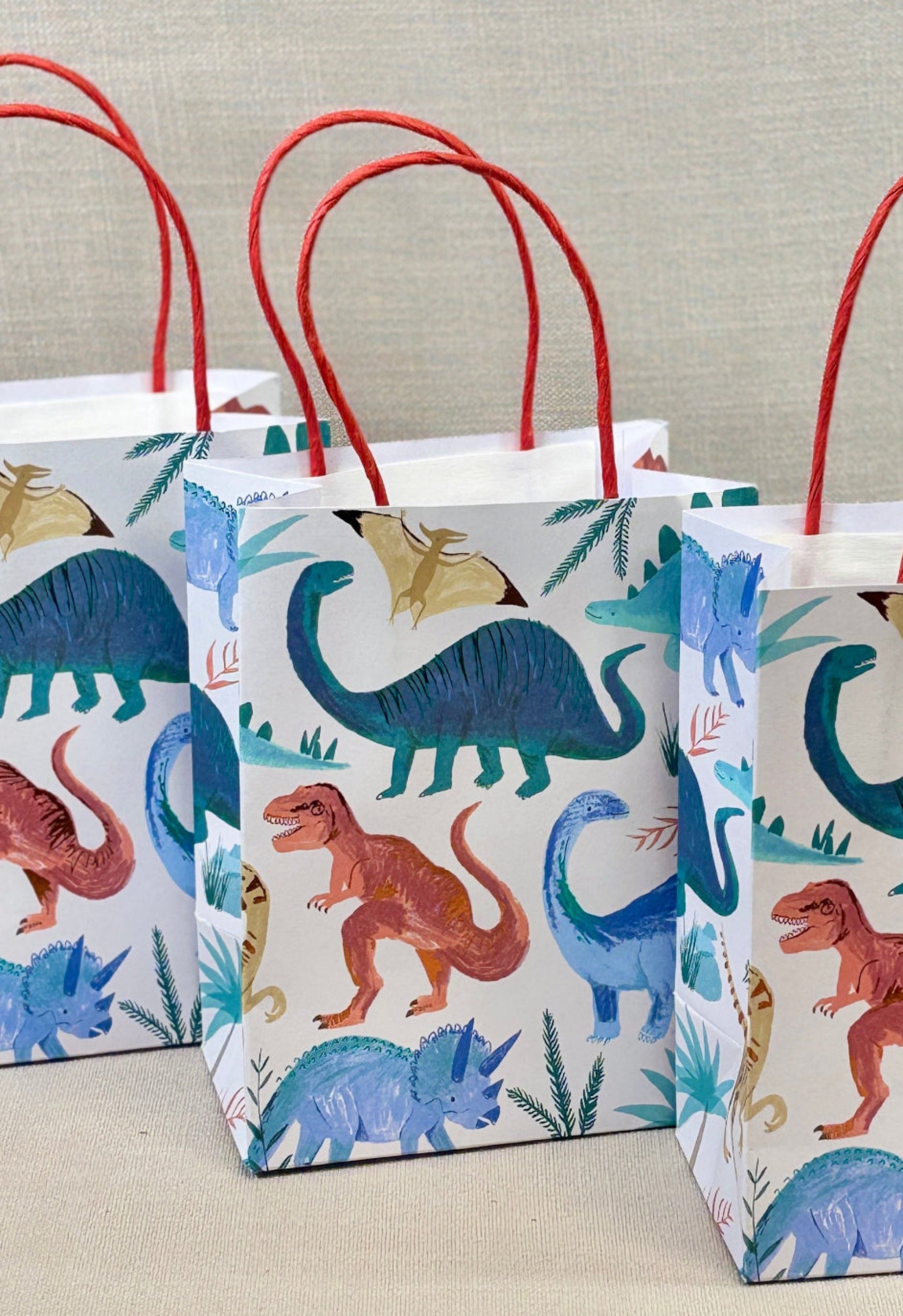 Three paper gift bags with dinosaur pattern on a neutral background