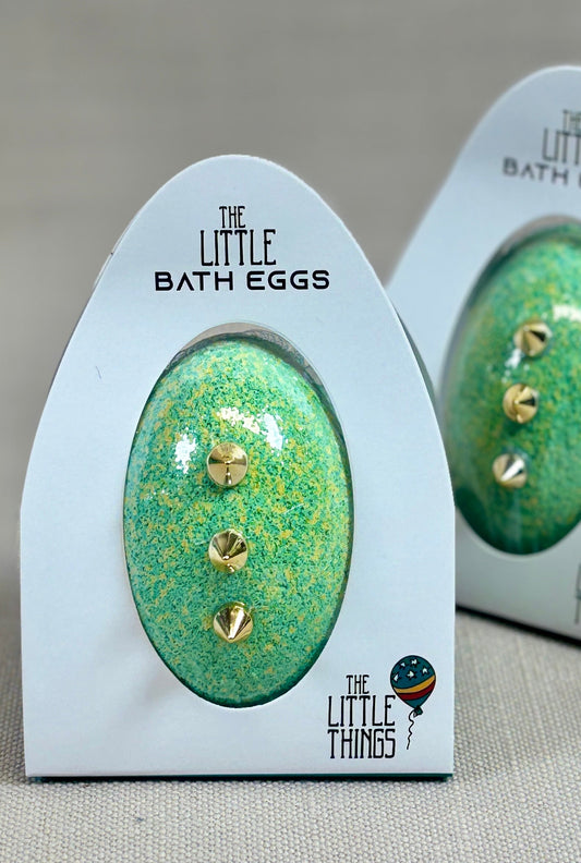 Two green bath eggs with gold accents in their packaging on a beige background