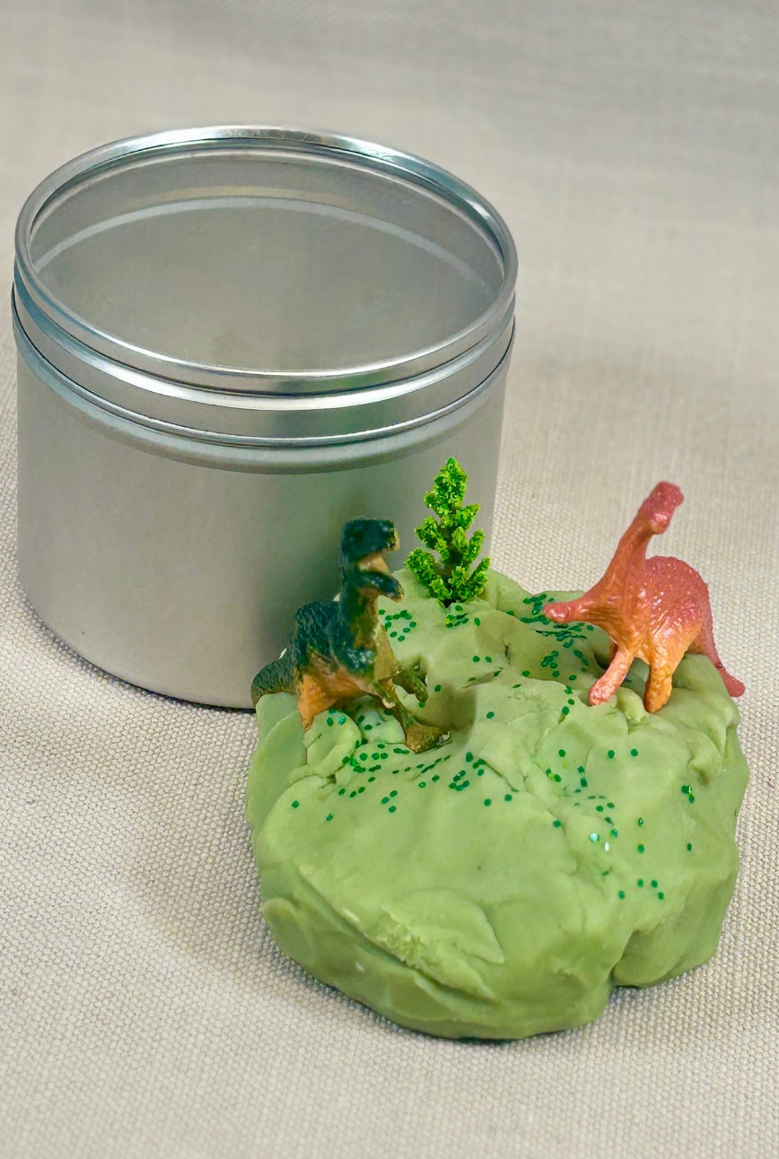 Green homemade playdoh with dinosaur figures next to a metal tin.