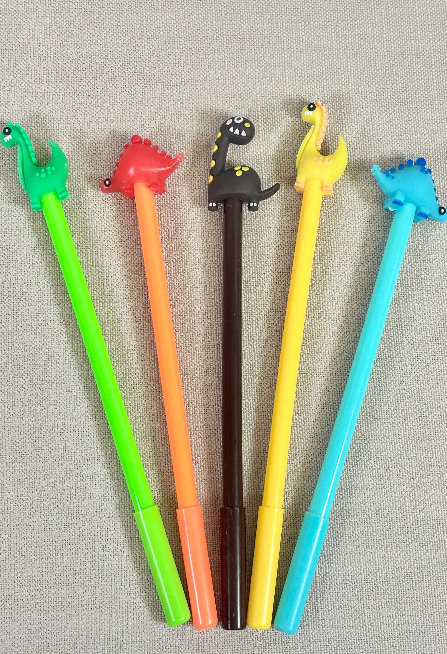 Five colorful pens with dinosaur-shaped tips on a textured gray background