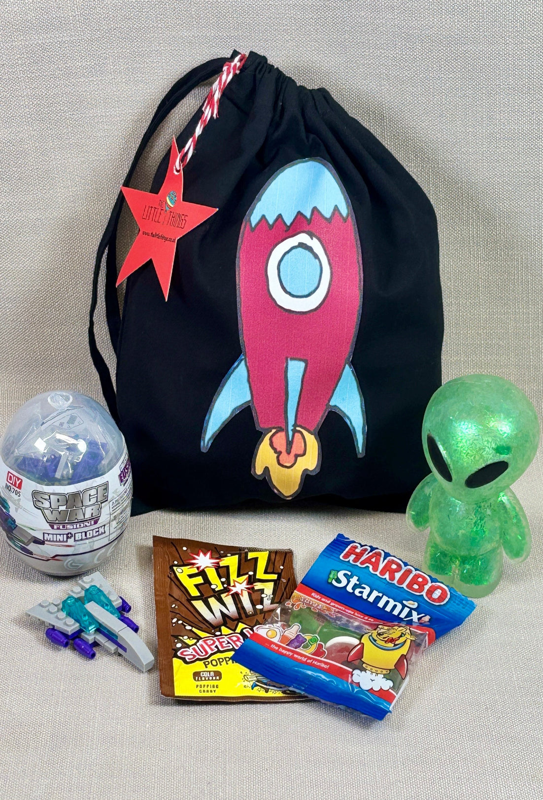 Black drawstring party bag with a rocket design, surrounded by small toys fillers, including a shish alien, space war mini bricks, fizz whizz and a star mix haribo.