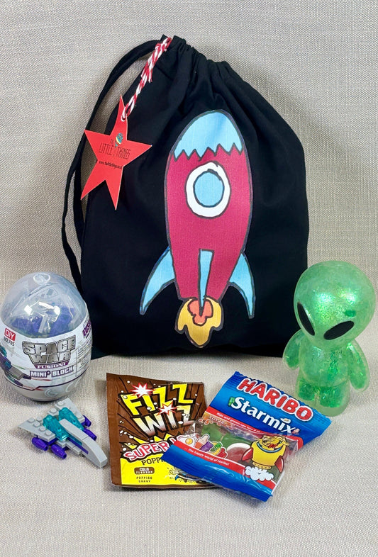 Black drawstring party bag with a rocket design, surrounded by small toys fillers, including a shish alien, space war mini bricks, fizz whizz and a star mix haribo.