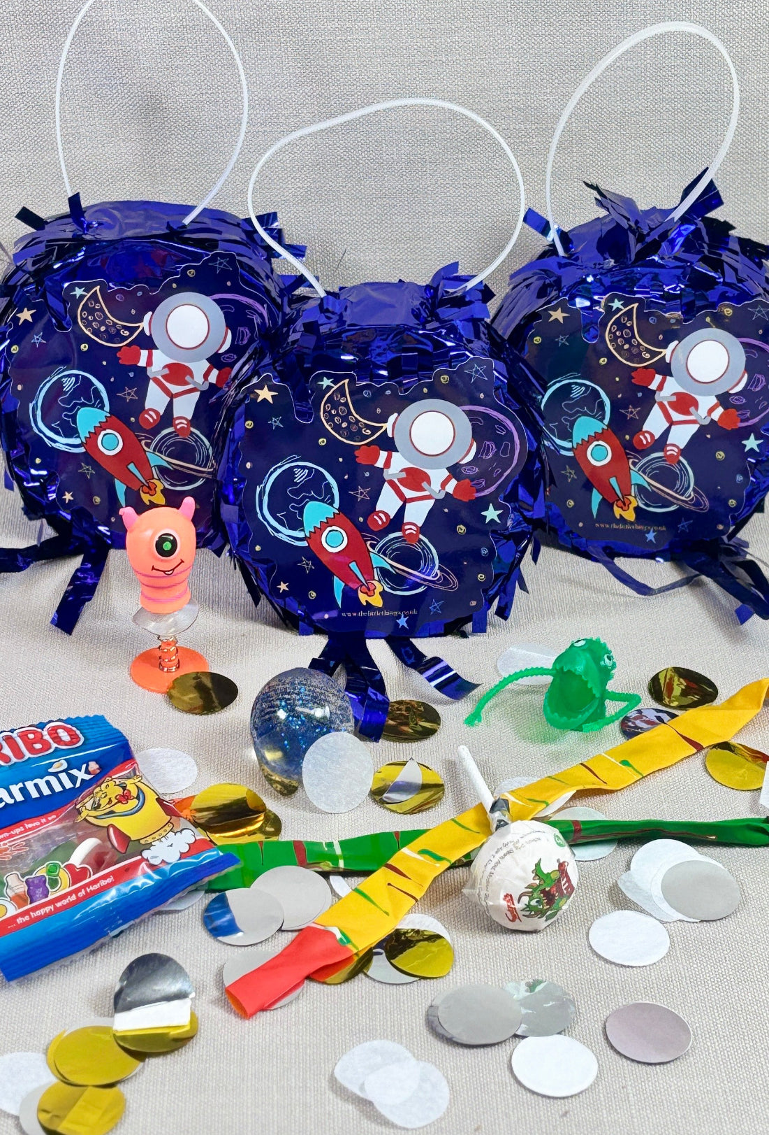 Decorative piñata items including blue bags with space-themed designs, colorful ribbons, and confetti on a gray surface.
