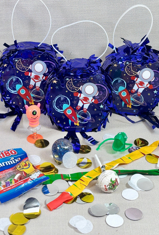 Decorative piñata items including blue bags with space-themed designs, colorful ribbons, and confetti on a gray surface.