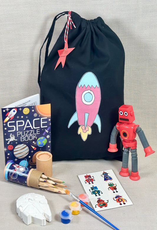 Space-themed children's activity set with a drawstring bag, puzzle book, robot toy, and art supplies on a gray background.