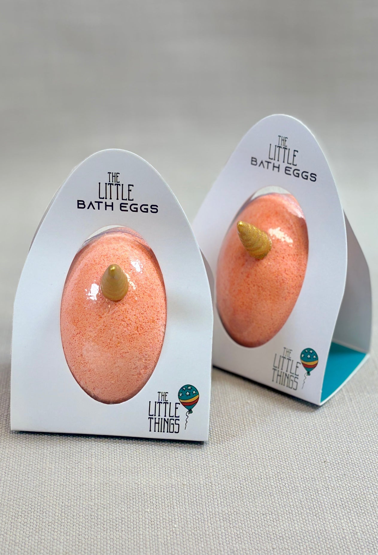 Two peach-colored bath bombs in packaging with 'The Little Things' branding on a neutral background.
