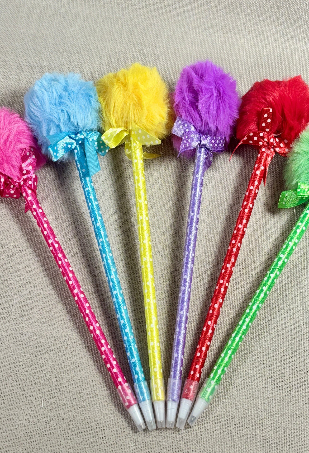 Six decorative fluffy pens in pink, blue, yellow, purple, red, and green, arranged in a fan shape on a neutral gray textured fabric background.