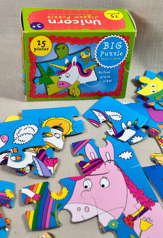 A children's jigsaw puzzle set with a colorful box labeled 'Unicorn Big Puzzle' and several loose puzzle pieces scattered in front on a neutral fabric surface.