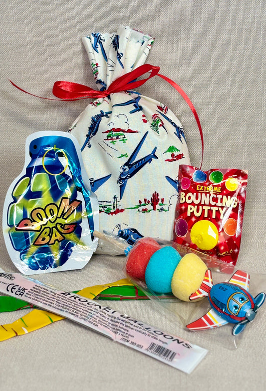 A gift set of retro toys and candy arranged on a beige linen fabric background. The set includes a white drawstring bag with a blue aoeroplan print, a  Plane sweet kabab, Bouncing Putty, Boom Bag, and 2x rocket balloons. 