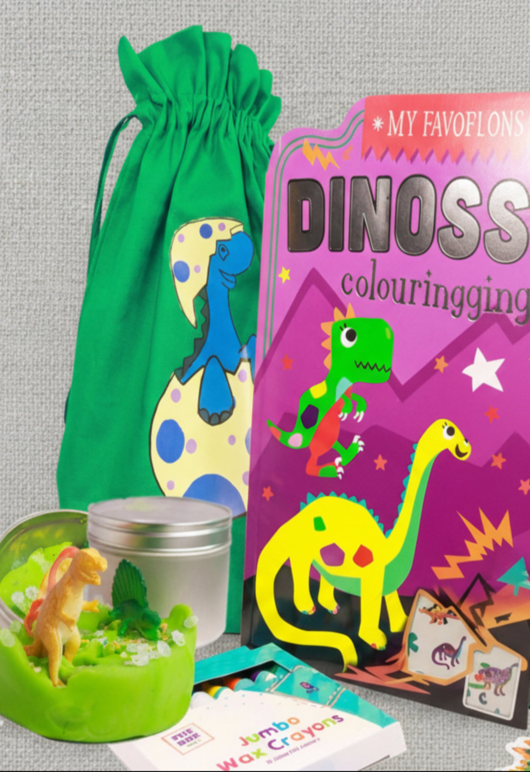 A large green fabric party bag with dinosaur print alongside a dinosaur-themed coloring book, playdough kit,  and jumbo crayons.